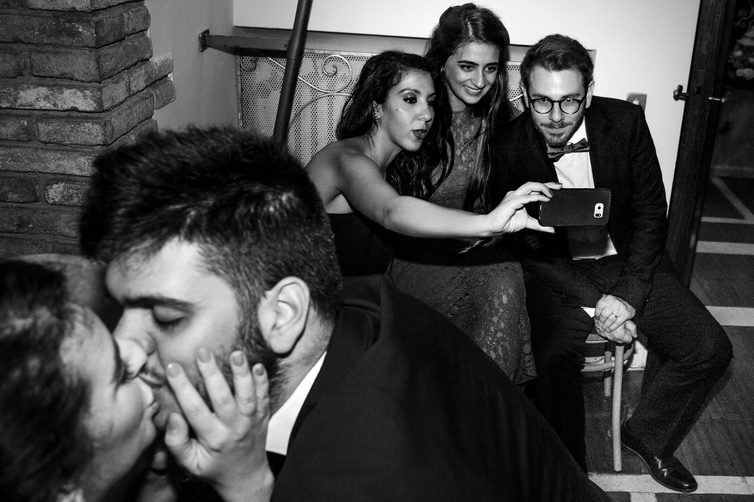 selfie at wedding while another couple kisses multi subject street photography black and white wedding photography