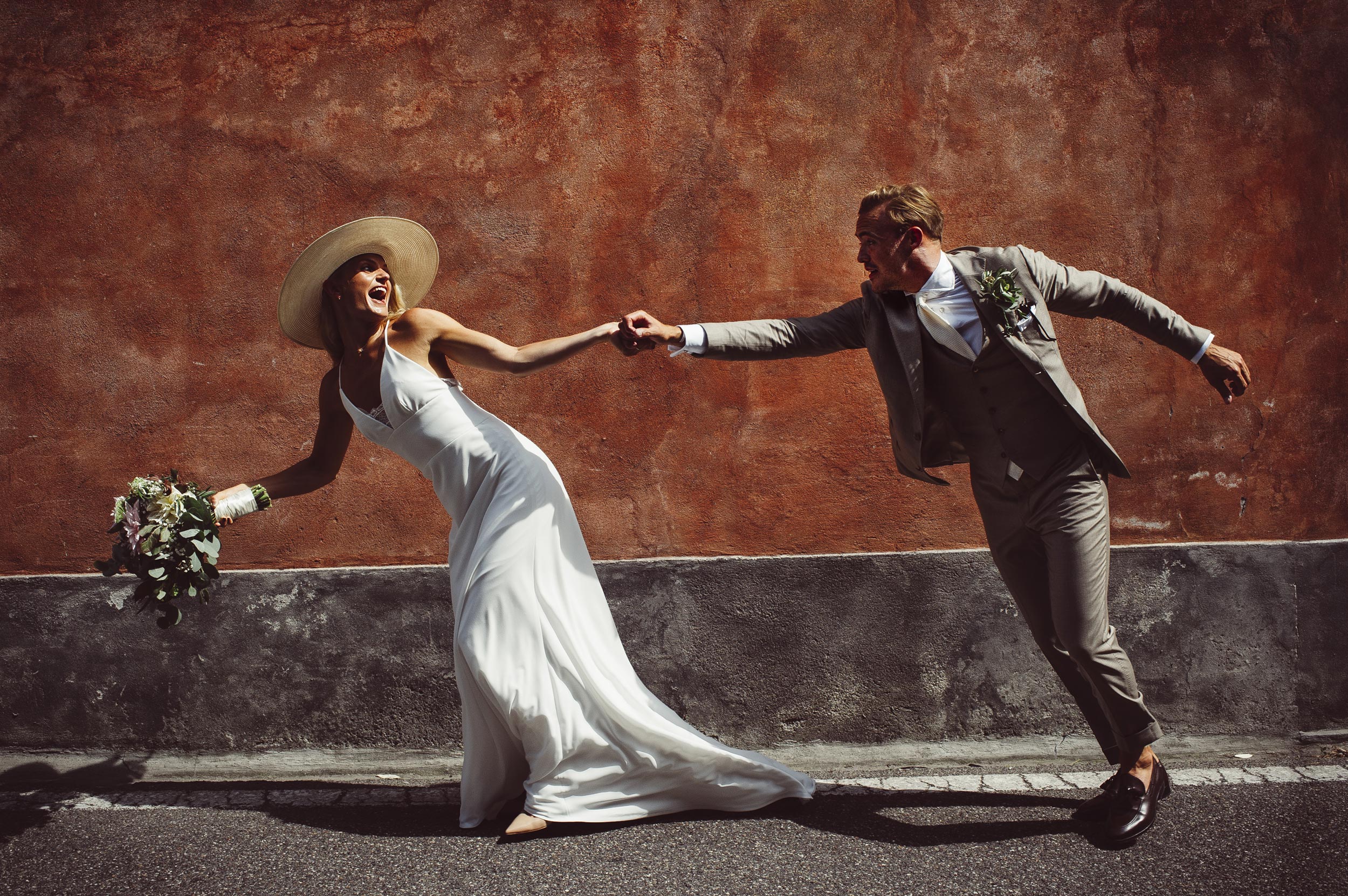 lake como wedding photographer Alessandro Avenali bride with hat grabs the groom running against a red wall